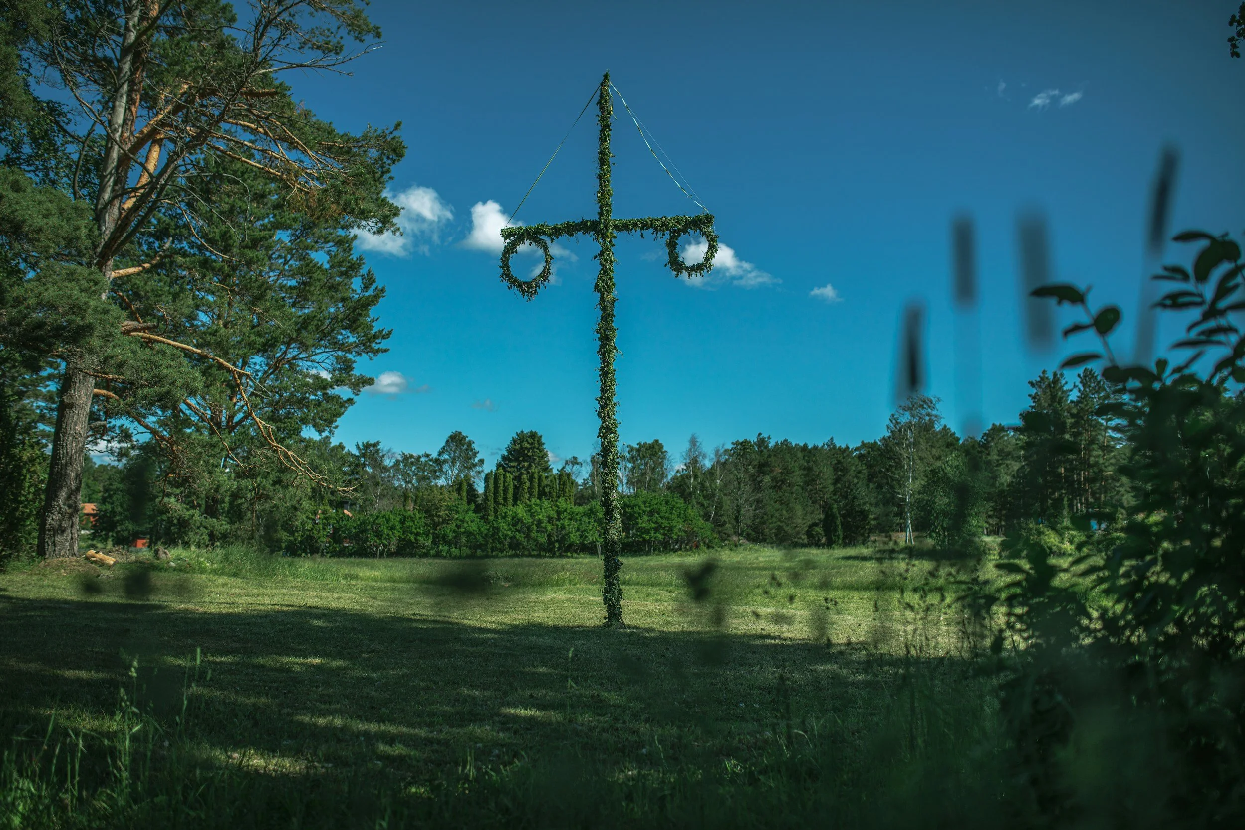A large cross-shaped structure covered in greenery and hanging wreaths, standing in an open grassy field with trees and a clear blue sky in the background.