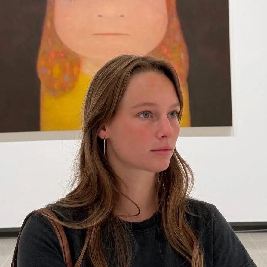 Young woman with long brown hair wearing a black shirt, standing in an art gallery with a colorful abstract painting in the background.