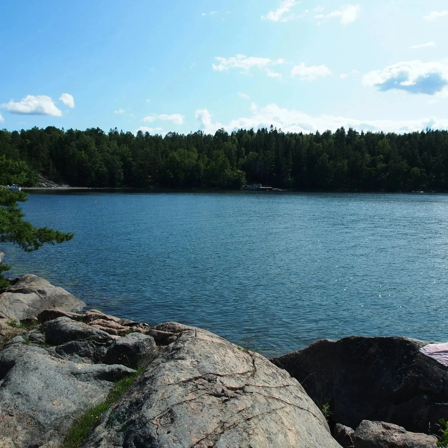 A peaceful lakeside scene with large rocks in the foreground, calm blue water, a dense forest on the far shore, and a partly cloudy sky overhead.
