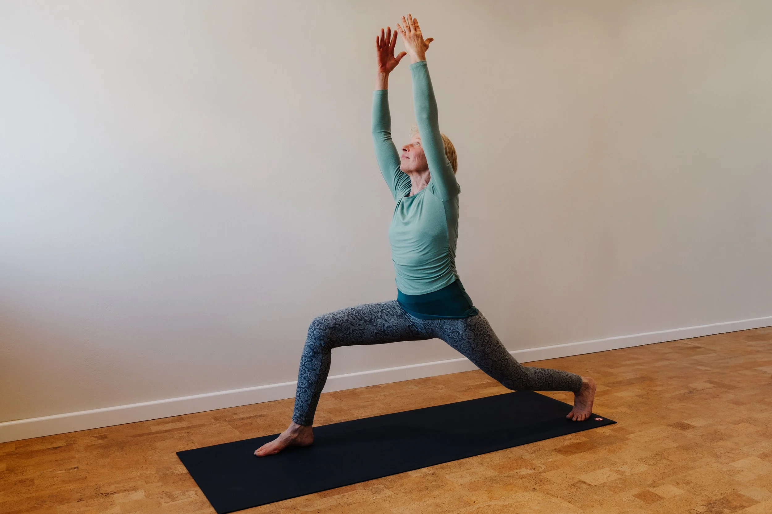 Eine Frau macht Yoga im Innenraum, steht in der Warrior-Position auf einem schwarzen Yogamatte. Sie trägt ein graues langärmeliges Oberteil und bunte Leggings mit Muster. Die Wand im Hintergrund ist beige, der Boden Holzdielen.