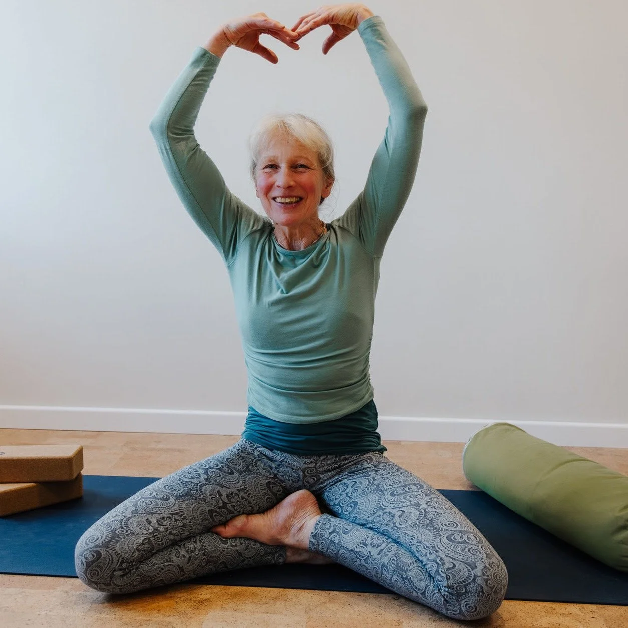 Eine ältere Frau macht Yoga im Sitzen, mit den Beinen verschränkt, und formt ein Herz mit den Händen über dem Kopf, vor einer weißen Wand.
