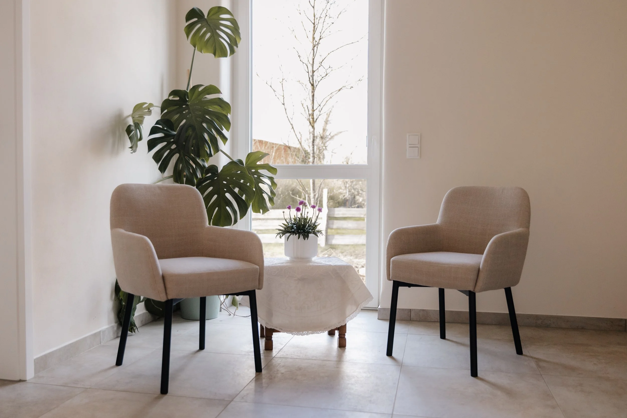 Zwei beige Stühle um einen kleinen, runden Tisch mit weißem Tuch, auf dem eine Topfpflanze mit lila Blumen steht, vor einer großen Fensterfront mit Blick nach draußen auf einen Baum und einen Garten. An der Wand rechts sind zwei Lichtschalter.