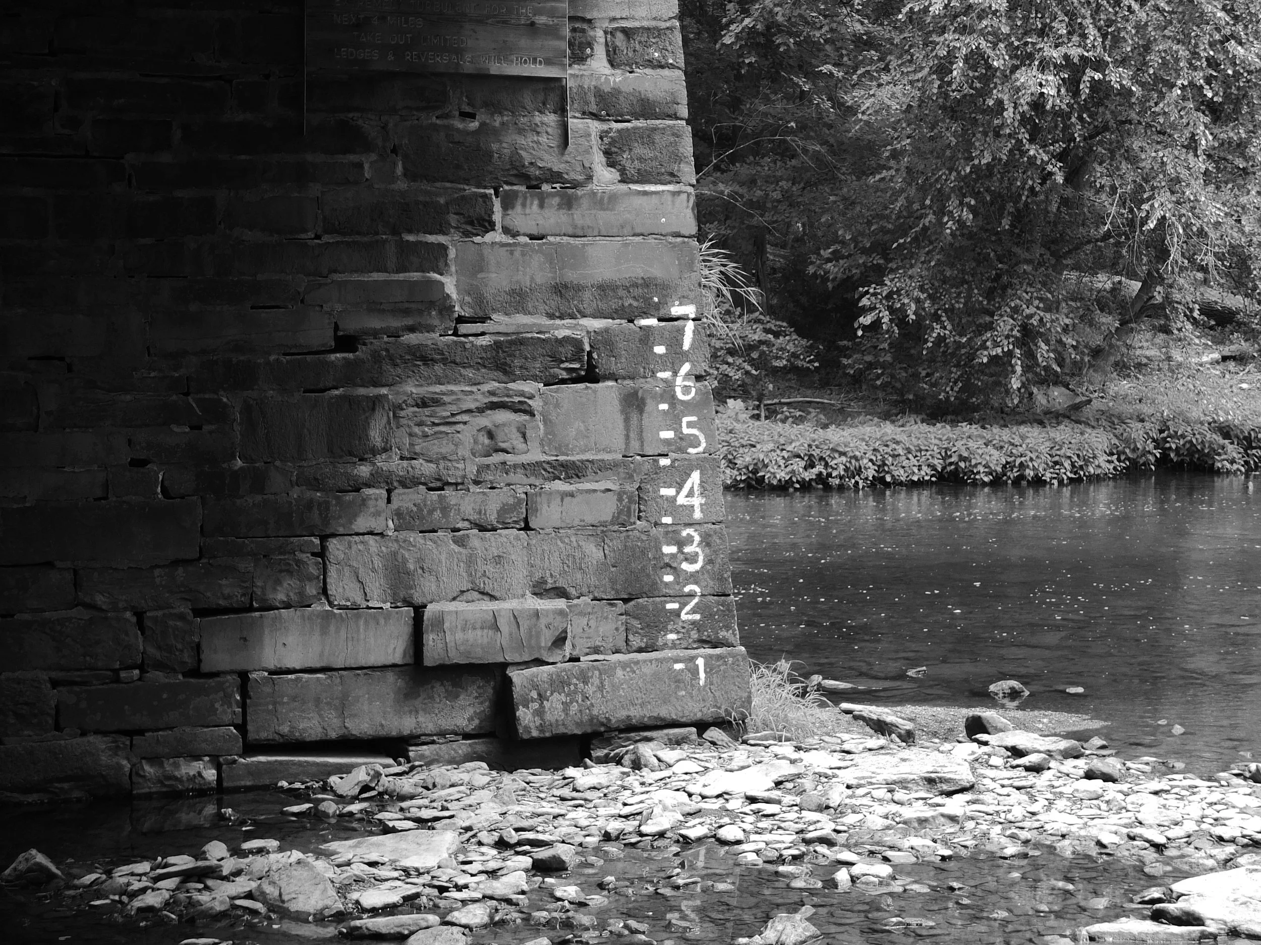 A black-and-white photo of a stone bridge over a creek with a measuring stick attached to the bridge's side, showing water levels, surrounded by trees and plants.