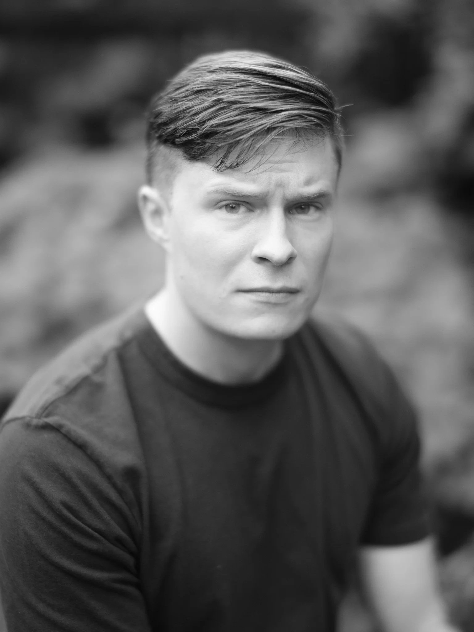 A black and white close-up portrait of a young man with short, styled hair and a serious expression, wearing a dark jacket.