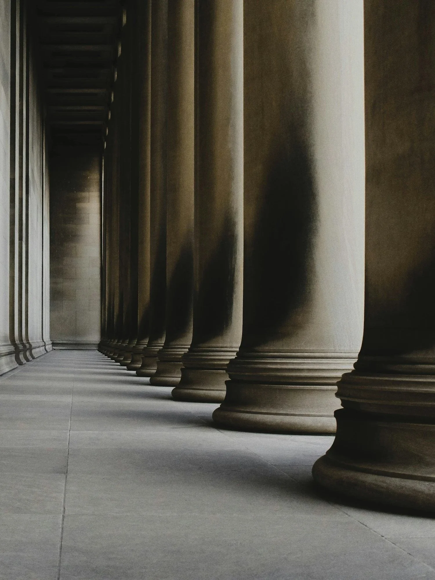 A row of large, classical stone columns in a building, with shadows cast on the floor.