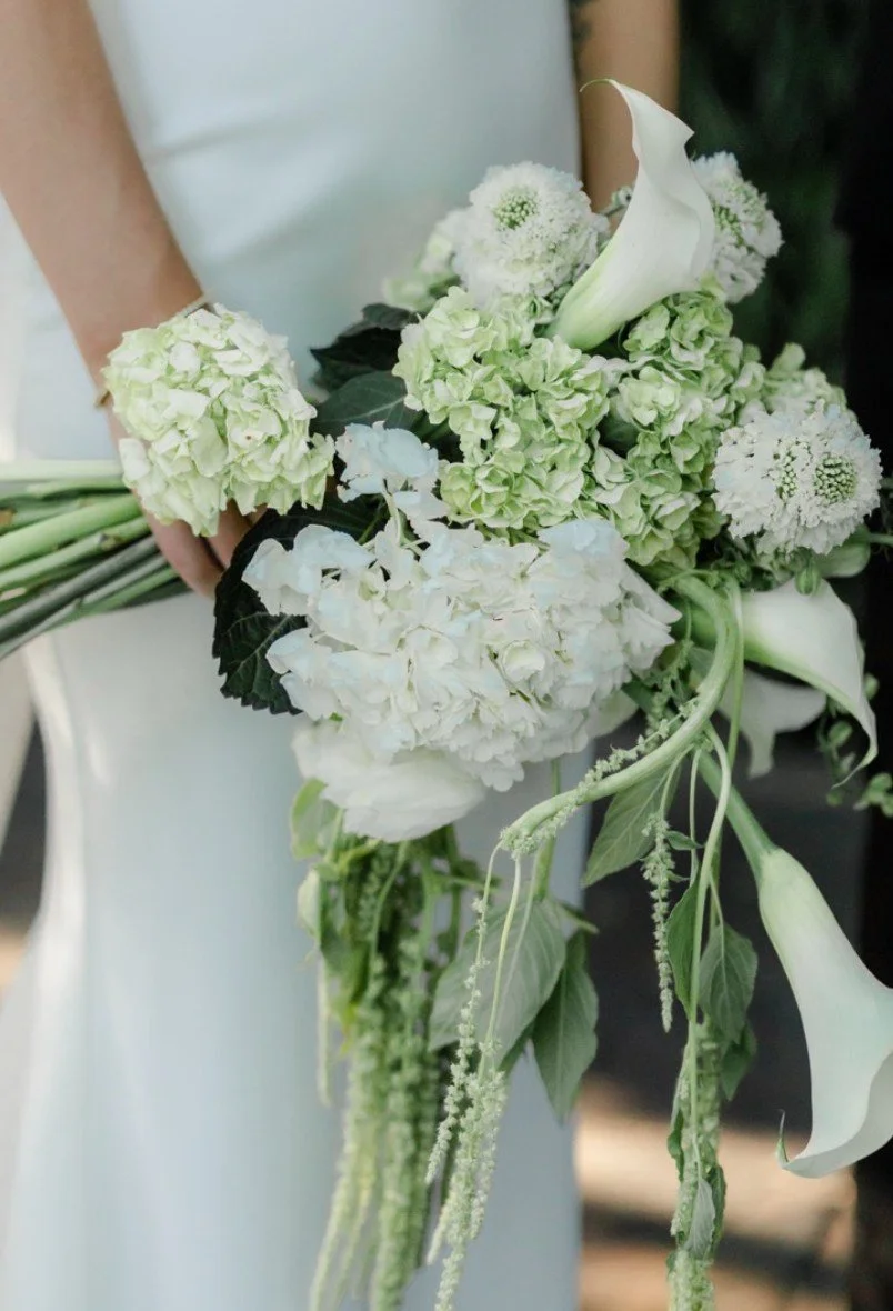 A person holding a cascading bouquet of white and pale green flowers, including calla lilies, hydrangeas, and various greenery.