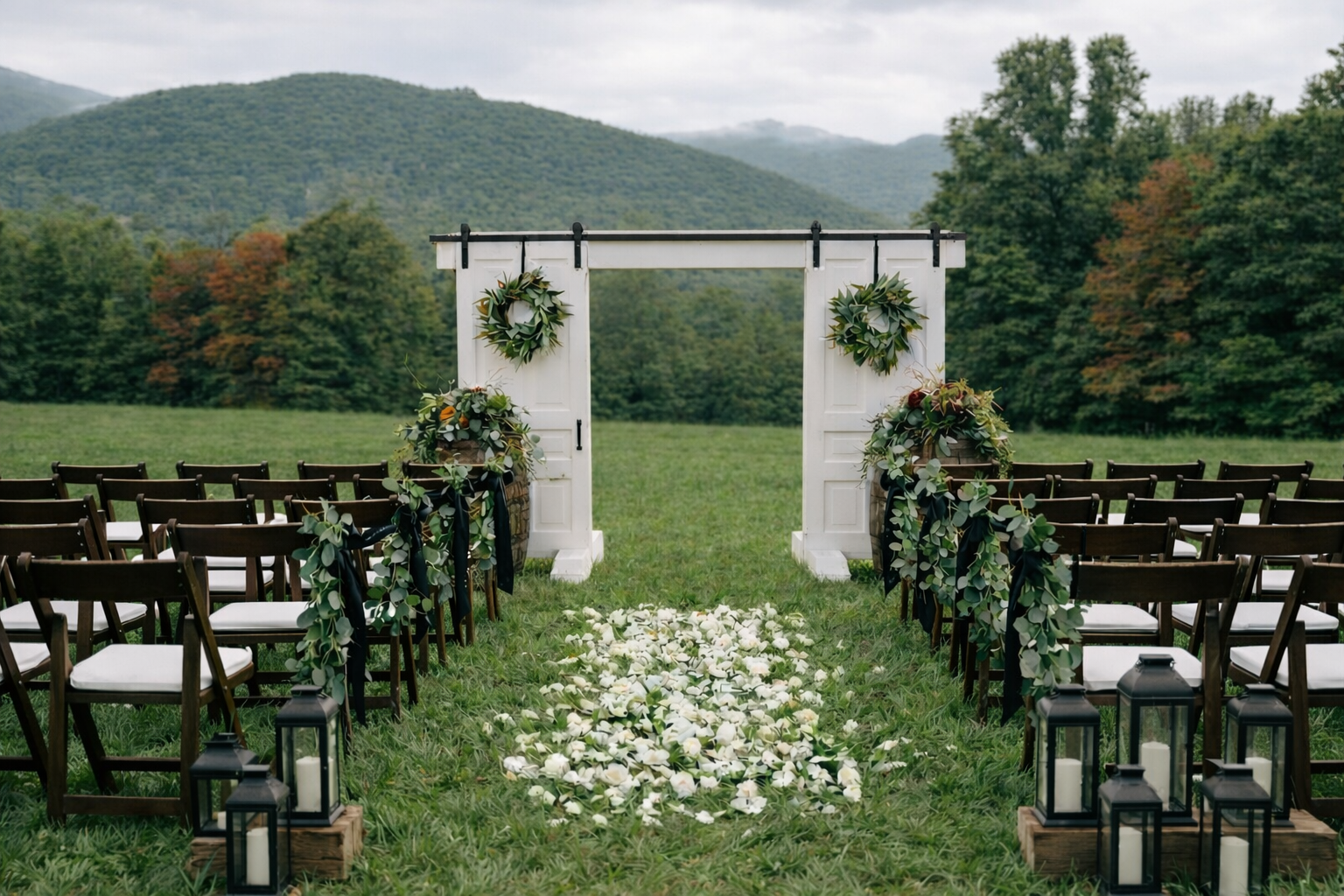 Outdoor wedding ceremony setup on a grassy field with chairs, floral decorations, lanterns, and a white wooden arch against a backdrop of trees and mountains.