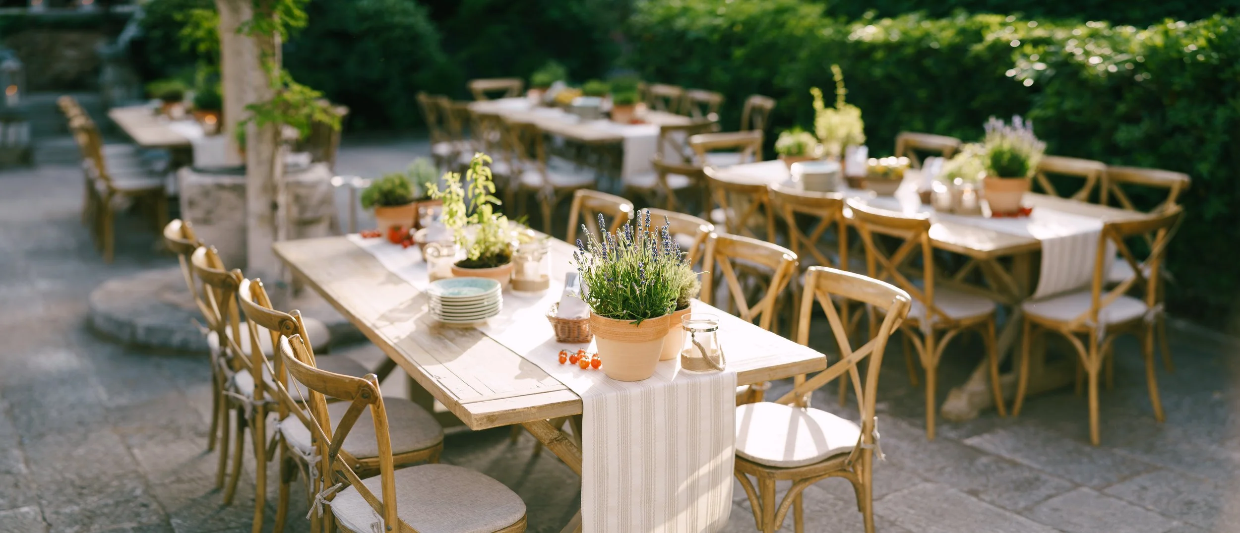Outdoor dining setup with wooden tables and chairs, decorated with potted plants, flowers, and white tablecloths, in a garden setting with trees and greenery.