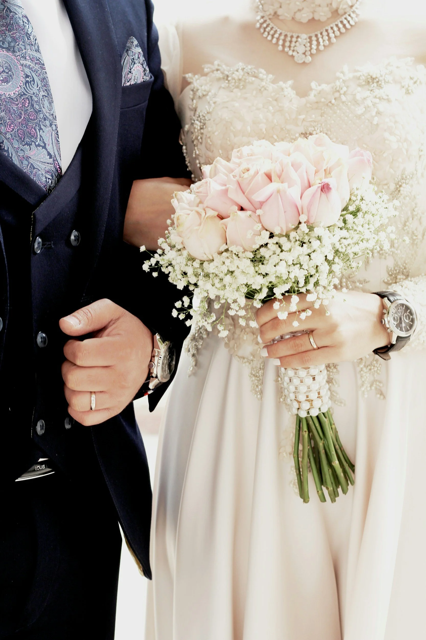 Close-up of a bride and groom, focusing on their hands and bouquet. The bride is holding a bouquet of pink roses and white baby's breath, wearing a wedding dress adorned with floral details. The groom is wearing a dark suit with a patterned tie, and both are wearing wedding rings.