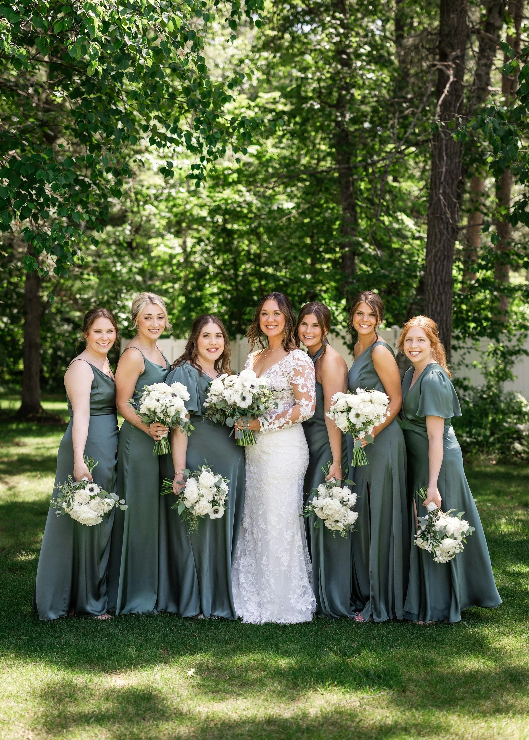 A bride and six bridesmaids standing outdoors on a grassy area beneath tall trees, all dressed in matching teal dresses, holding bouquets of white flowers, smiling at the camera.