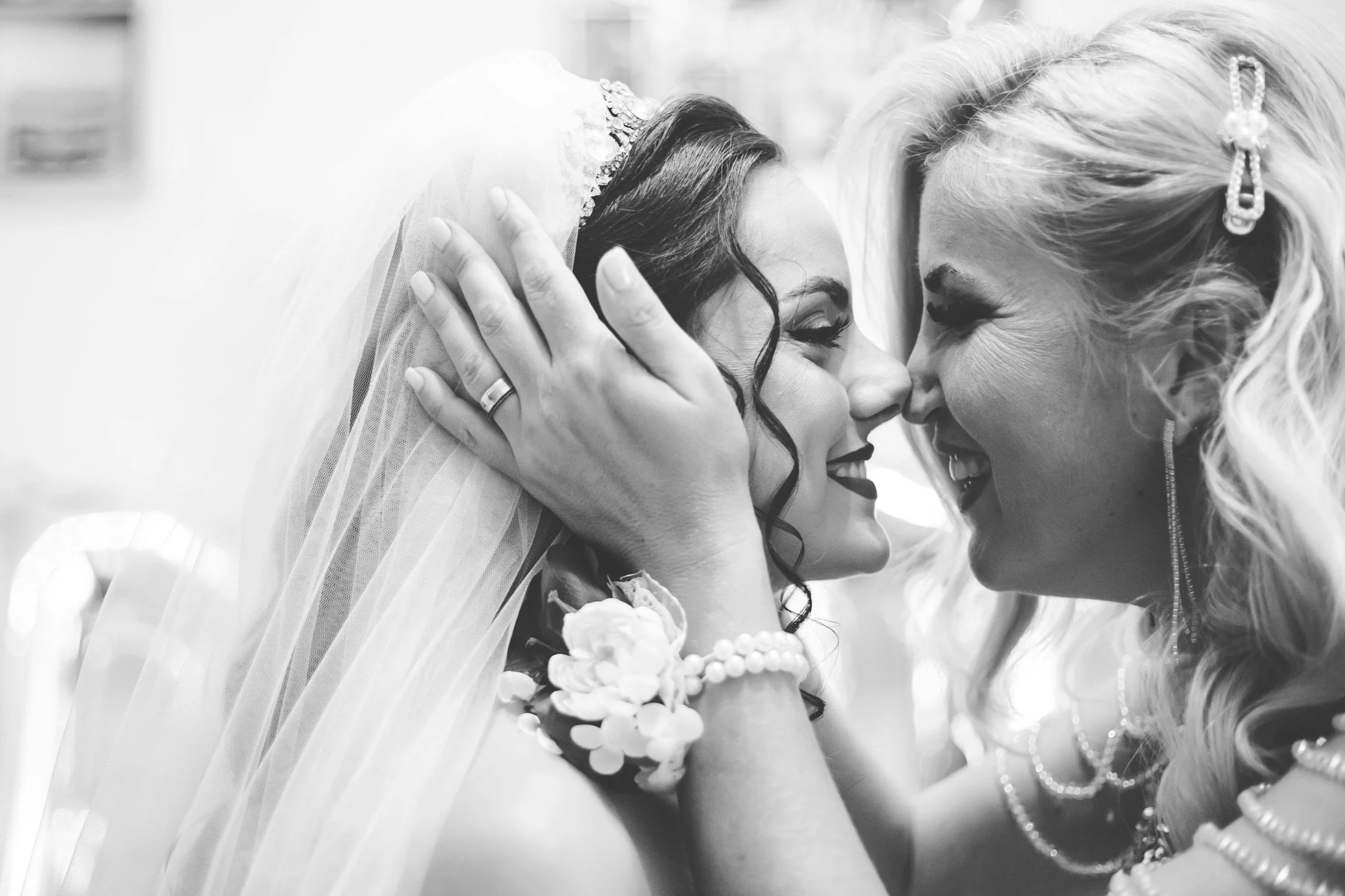 Black and white photo of two women touching foreheads and smiling, one in a wedding veil and crown, and the other with styled hair and jewelry, showing affection and joy.