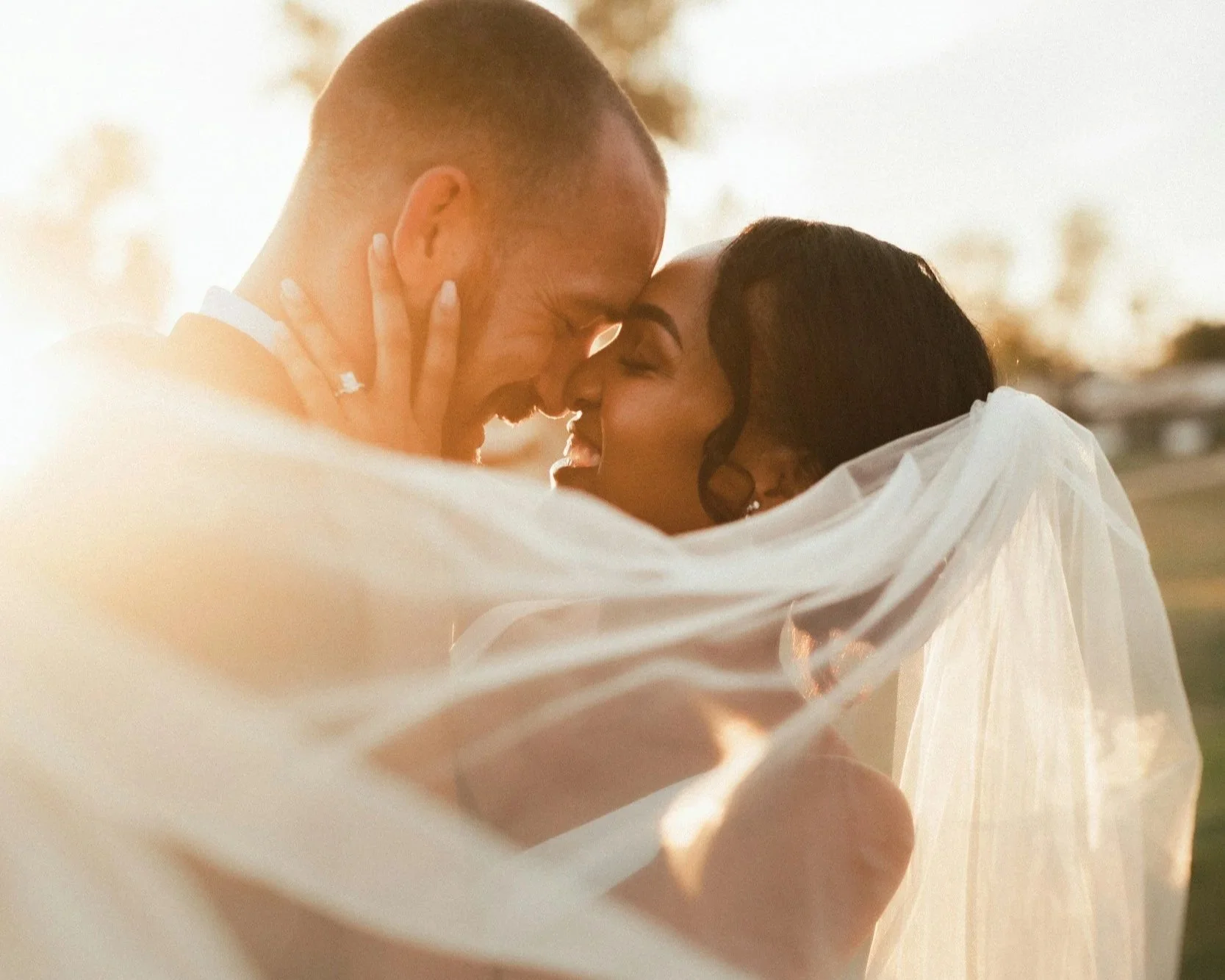 A bride and groom embrace outdoors during sunset, touching foreheads and smiling with eyes closed, wrapped in a white veil.