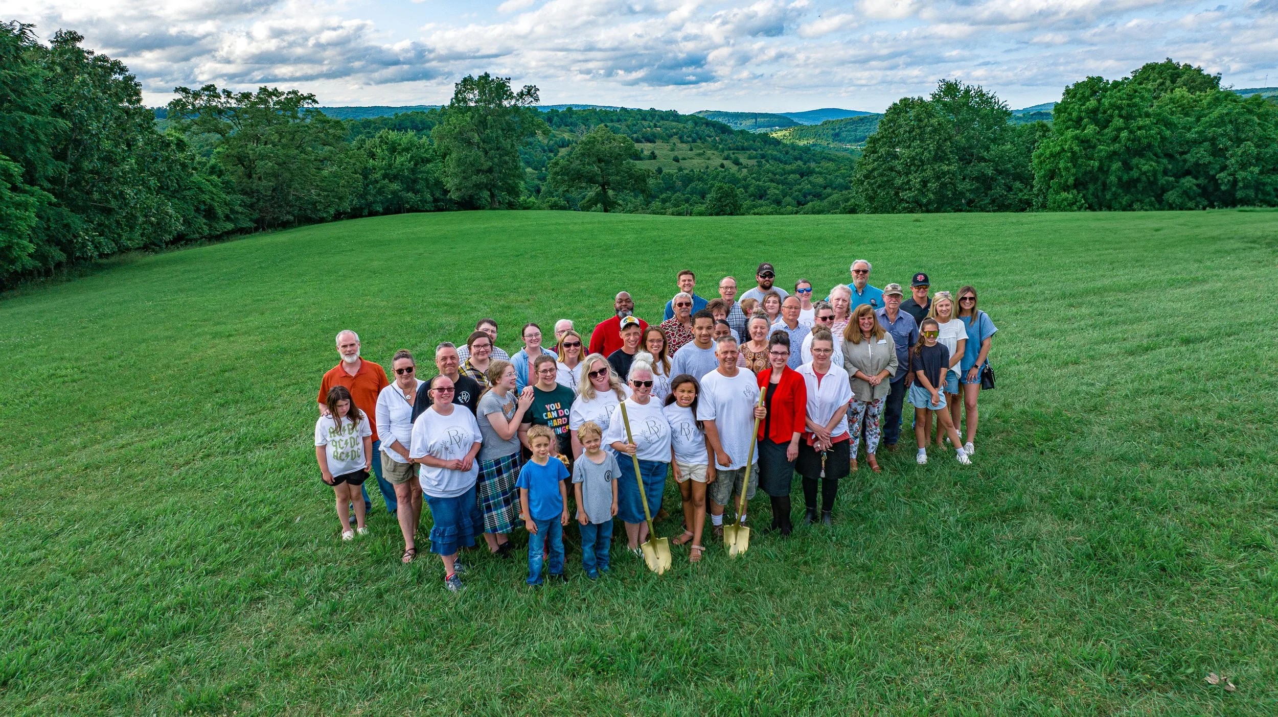 A large group of people standing on a green grassy field with trees and hills in the background, under a partly cloudy sky.