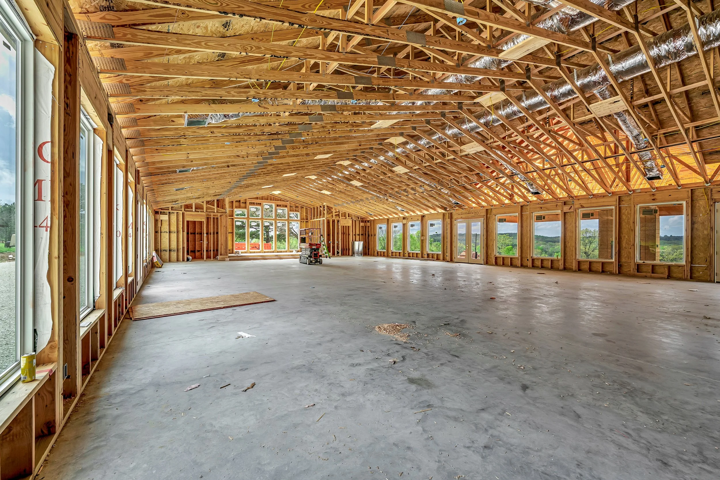 Construction site of a large wooden house with exposed framing, windows, and open space inside, showing a view of green trees and a blue sky outside.