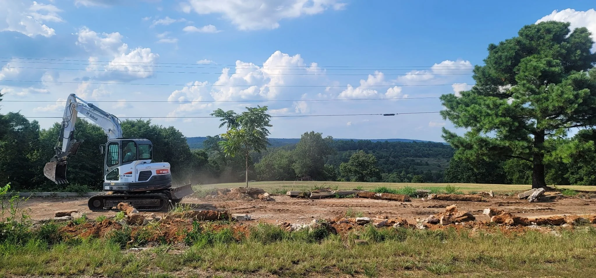 A small excavator working on clearing land with trees and a scenic hillside in the background under a partly cloudy sky.