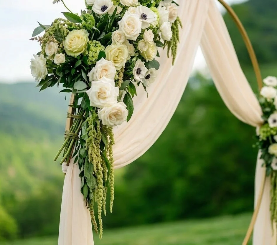 Floral wedding arch with white roses, anemones, and greenery, draped with cream fabric, set outdoors with a green landscape background.