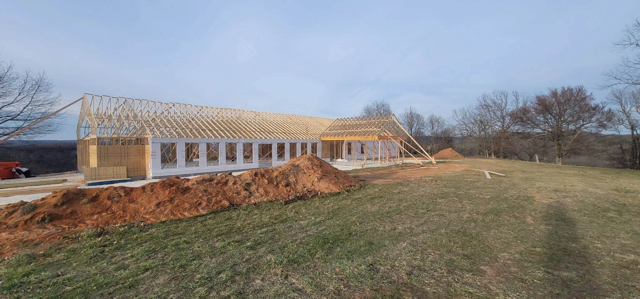 Construction site of a house with visible wooden framework and partially built walls, surrounded by dirt and grass, with trees in the background.