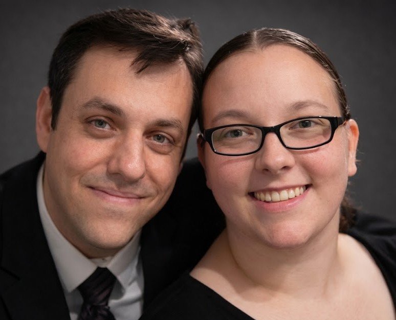 Close-up of a smiling man and woman with dark hair, wearing formal attire, posing together against a dark background.