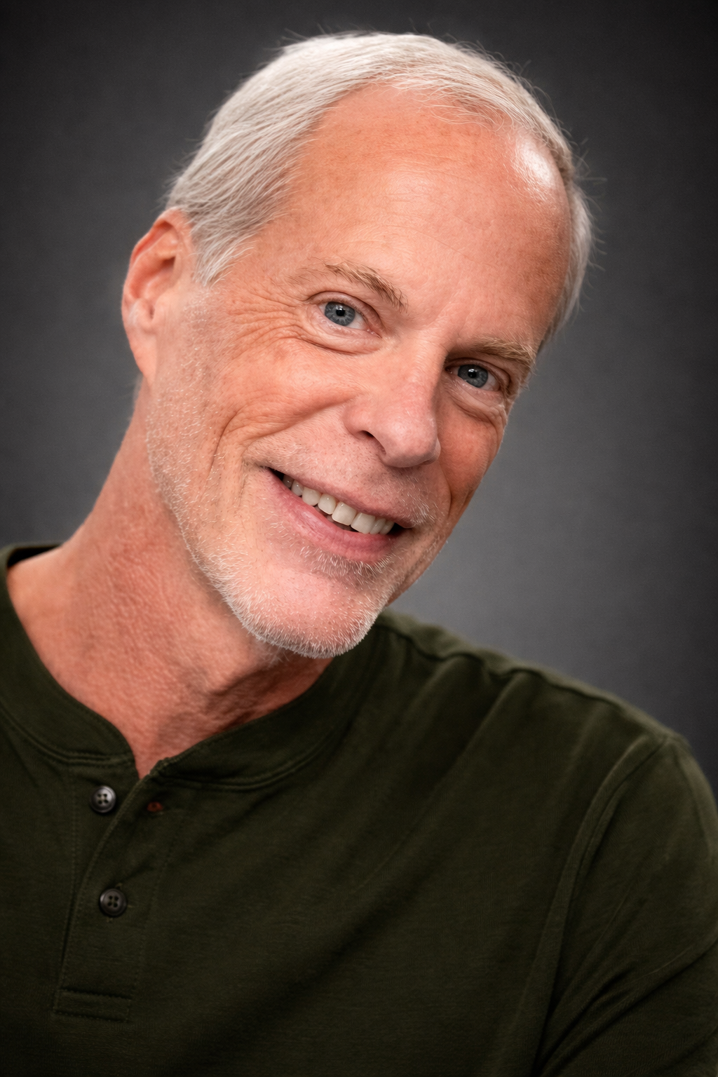 A smiling middle-aged man with short gray hair and blue eyes, wearing a dark green shirt, against a dark gray background.