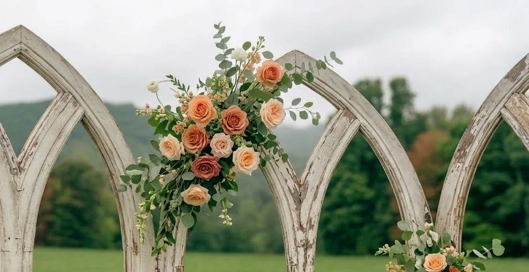 Floral arrangement of peach, blush, and deep pink roses with green foliage on an arched wooden trellis outdoors.
