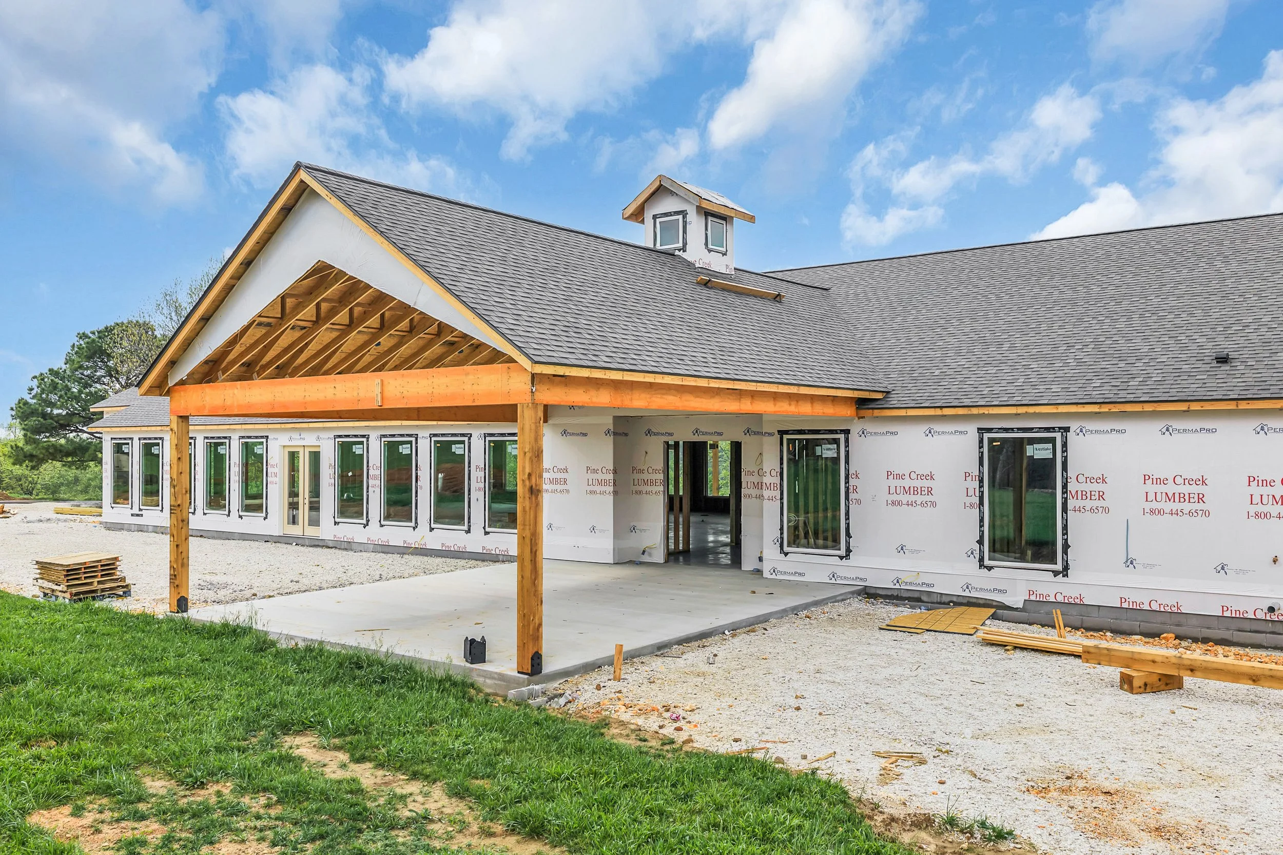 House under construction with a partially finished exterior, wooden porch support beams, and a gabled roof with a small cupola. The exterior walls are wrapped in building material, and multiple windows are installed. The surrounding area has a concrete pad and gravel, with some grass in the foreground and a partly cloudy blue sky overhead.