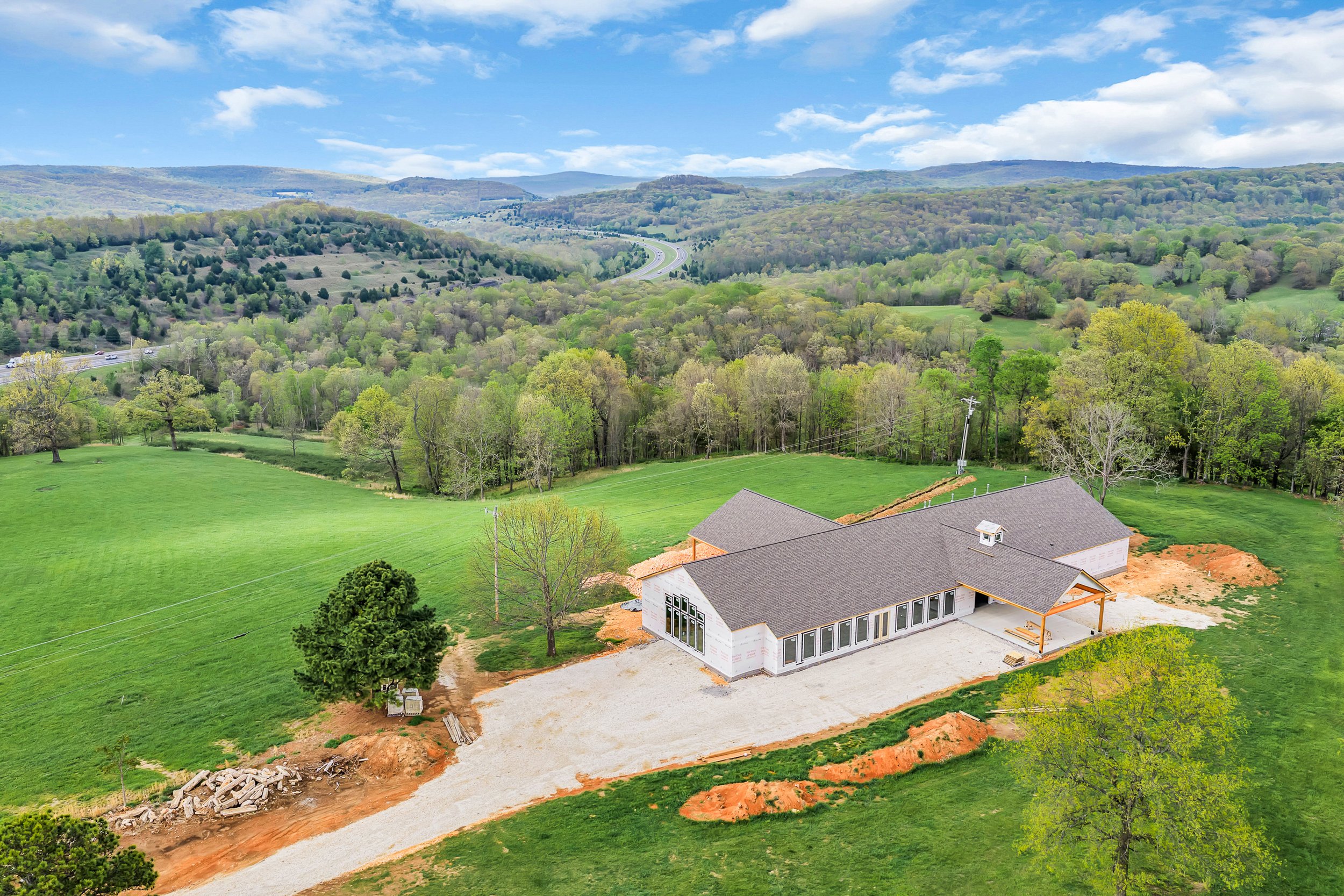 A house under construction in a green landscape with trees, rolling hills, a winding road, and a partly cloudy sky.