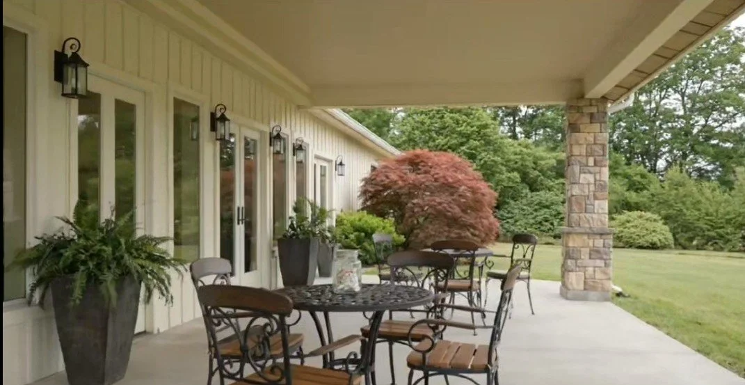 Covered outdoor patio with round metal table and chairs, potted plants, a reddish maple tree, and a large brick pillar, with a grassy yard and trees in the background.