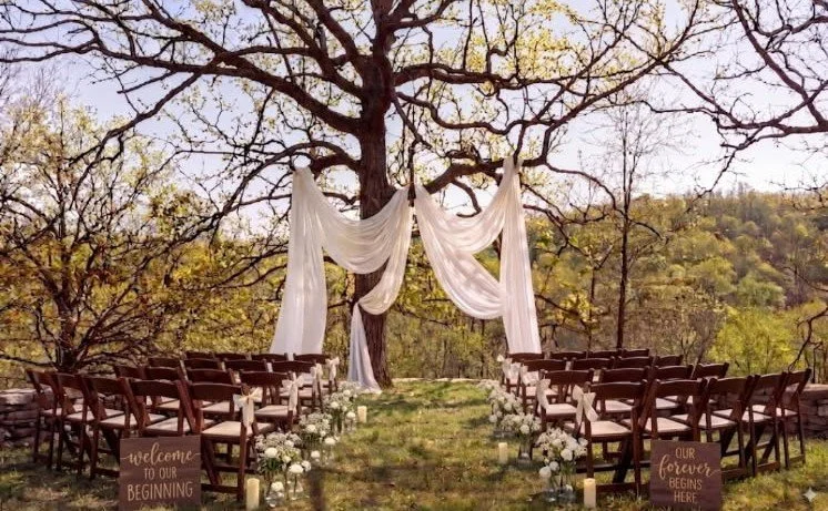Outdoor wedding ceremony setup with chairs, white floral decorations, and flowing white fabric draped from a tree, with scenic natural background and signs reading "Welcome to our beginning" and "Our forever begins here."