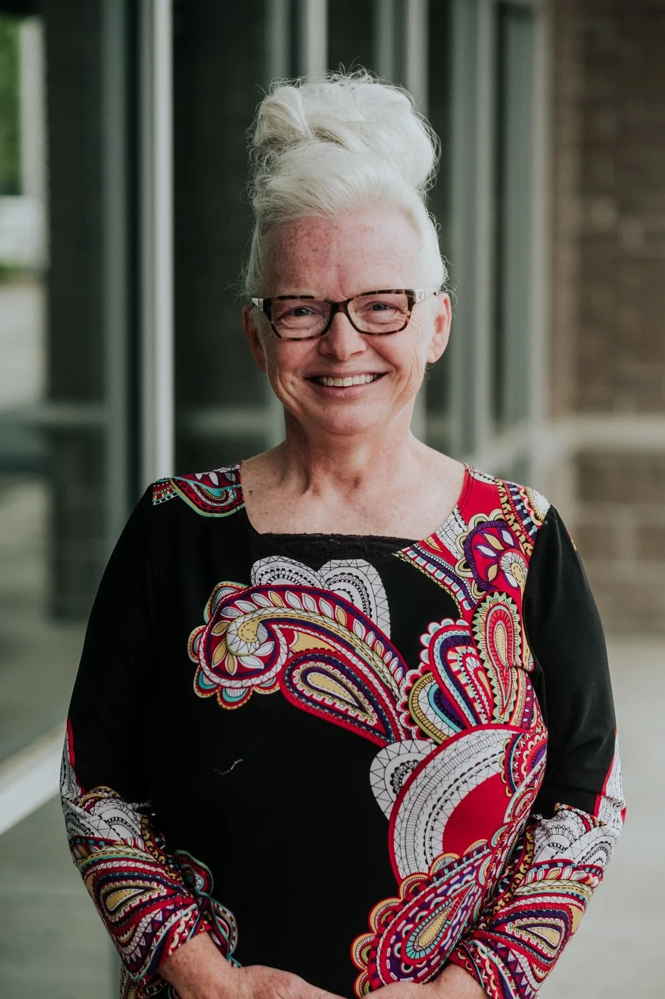 An elderly woman with white hair styled in a tall updo wearing glasses and a colorful paisley patterned dress, smiling outdoors.