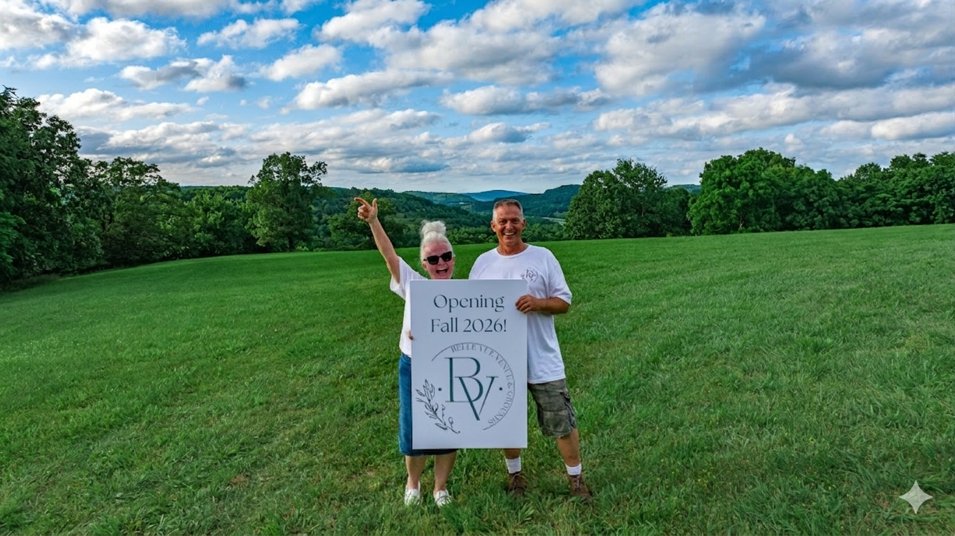 A man and woman standing in a green grassy field holding a sign that says 'Opening Fall 2026!' The woman is wearing sunglasses and a white shirt, and the man is wearing a white shirt and shorts. The sky is partly cloudy with scenic hills and trees in the background.