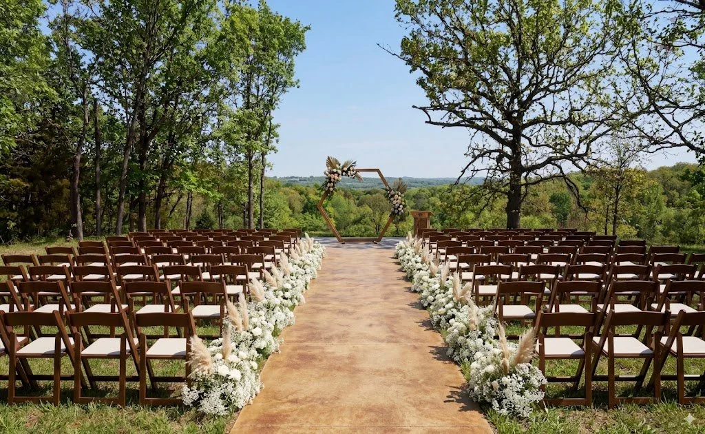 An outdoor wedding ceremony setup with rows of wooden chairs on either side of a wooden aisle, decorated with white flowers and pampas grass, leading to a hexagonal floral arch in a natural green setting with trees and a scenic view.