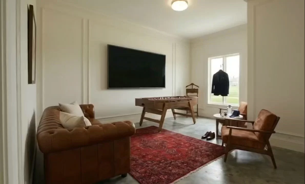 Living room with a large wall-mounted TV, leather couch with pillows, a vintage armchair, a foosball table, a side table with drinks and a phone, a window with a black coat hanging outside, and a red patterned rug on the floor.