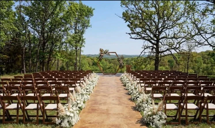 Outdoor wedding ceremony setup with rows of wooden chairs facing a floral arch on a lawn surrounded by trees.