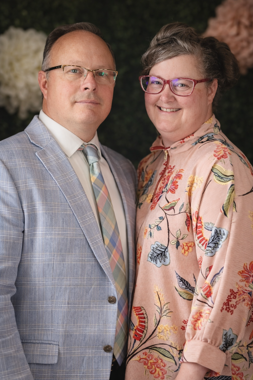 Portrait of a smiling woman with glasses, gray hair styled in a bun, wearing a floral top, against a dark background.