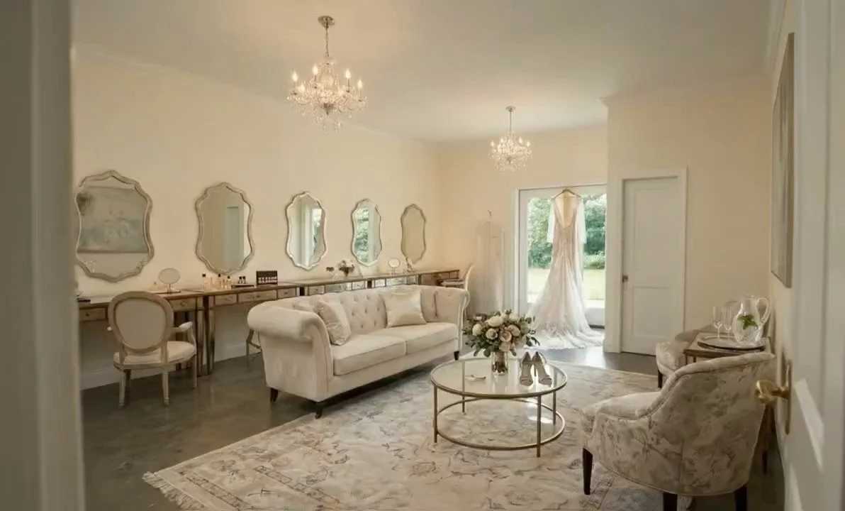 Elegant living room with white walls, a white sofa, floral armchairs, a round glass-top coffee table with a flower arrangement, and a long sideboard with mirrors and lamps, illuminated by two chandeliers.