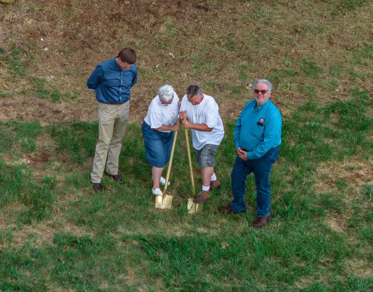 Four people standing outdoors on grass, two of them digging with shovels, one wearing a blue jacket, and a woman with glasses in a white shirt, while a man in a white shirt watches.