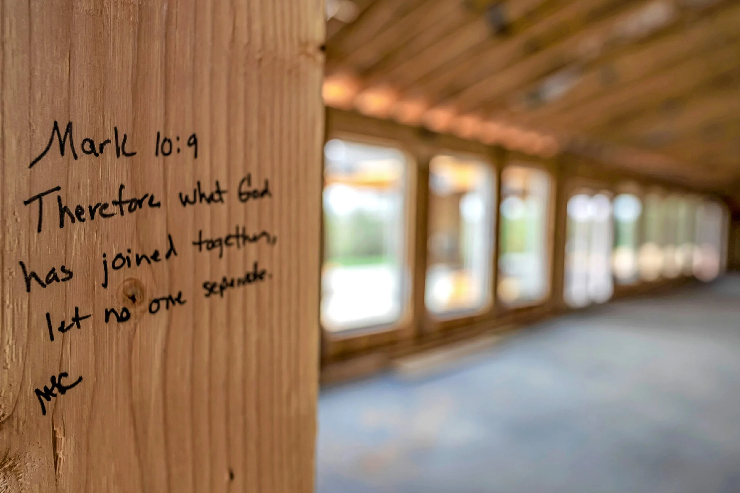 Close-up of a wooden wall with handwritten Bible verse referencing Mark 10:9, with blurred windows in the background.
