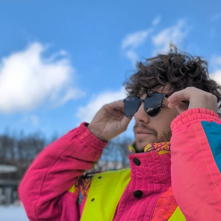 A man with curly hair donning sunglasses, about to hit the mountain in a vivid and extremely cool ski suit he bought in a second hand shop in Japan for only 6000 yen.