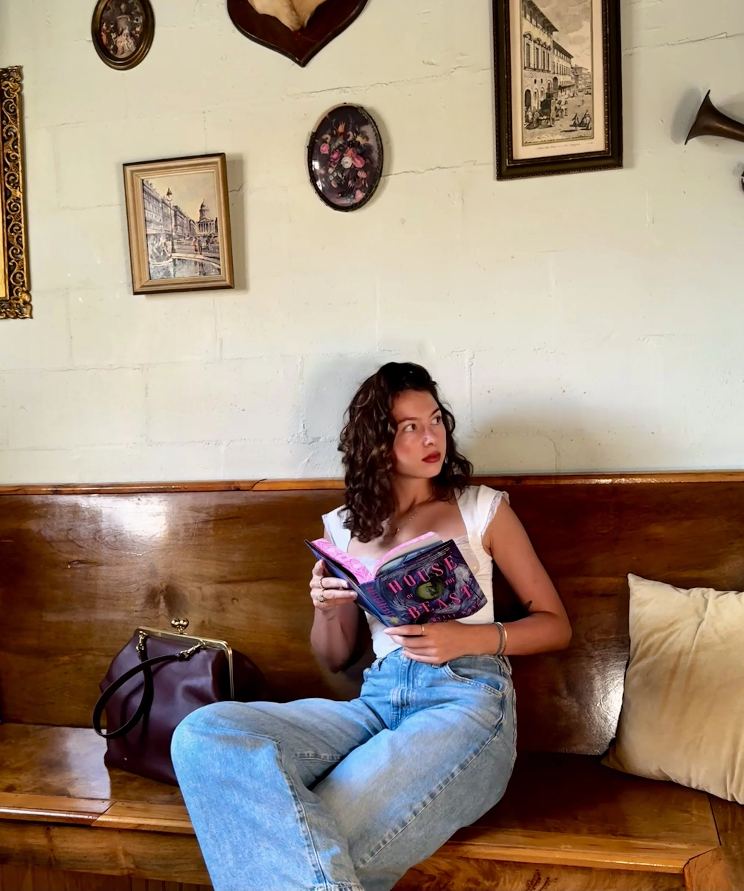 A young woman with curly hair sitting on a wooden bench, holding a colorful book titled 'House of Beast,' with a brown handbag beside her, in a vintage-styled room with framed pictures and art on the walls.