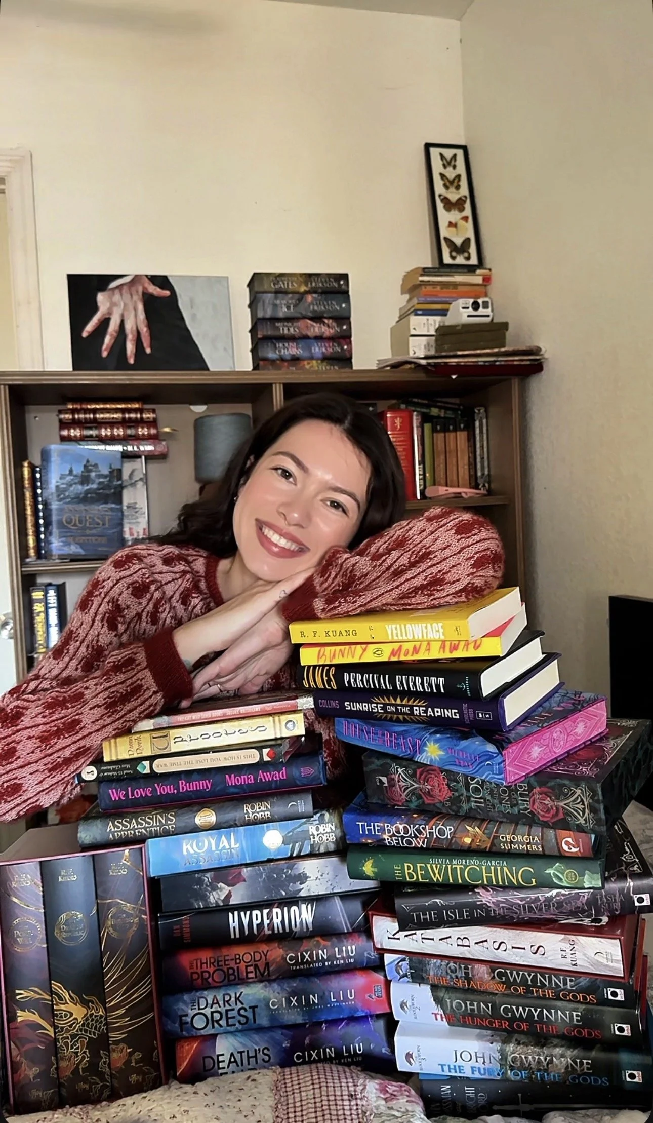 A woman with dark hair and a red patterned sweater lies resting her head on her crossed arms, beside a large stack of colorful books. The background shows a wooden bookshelf with more books and decorative items.