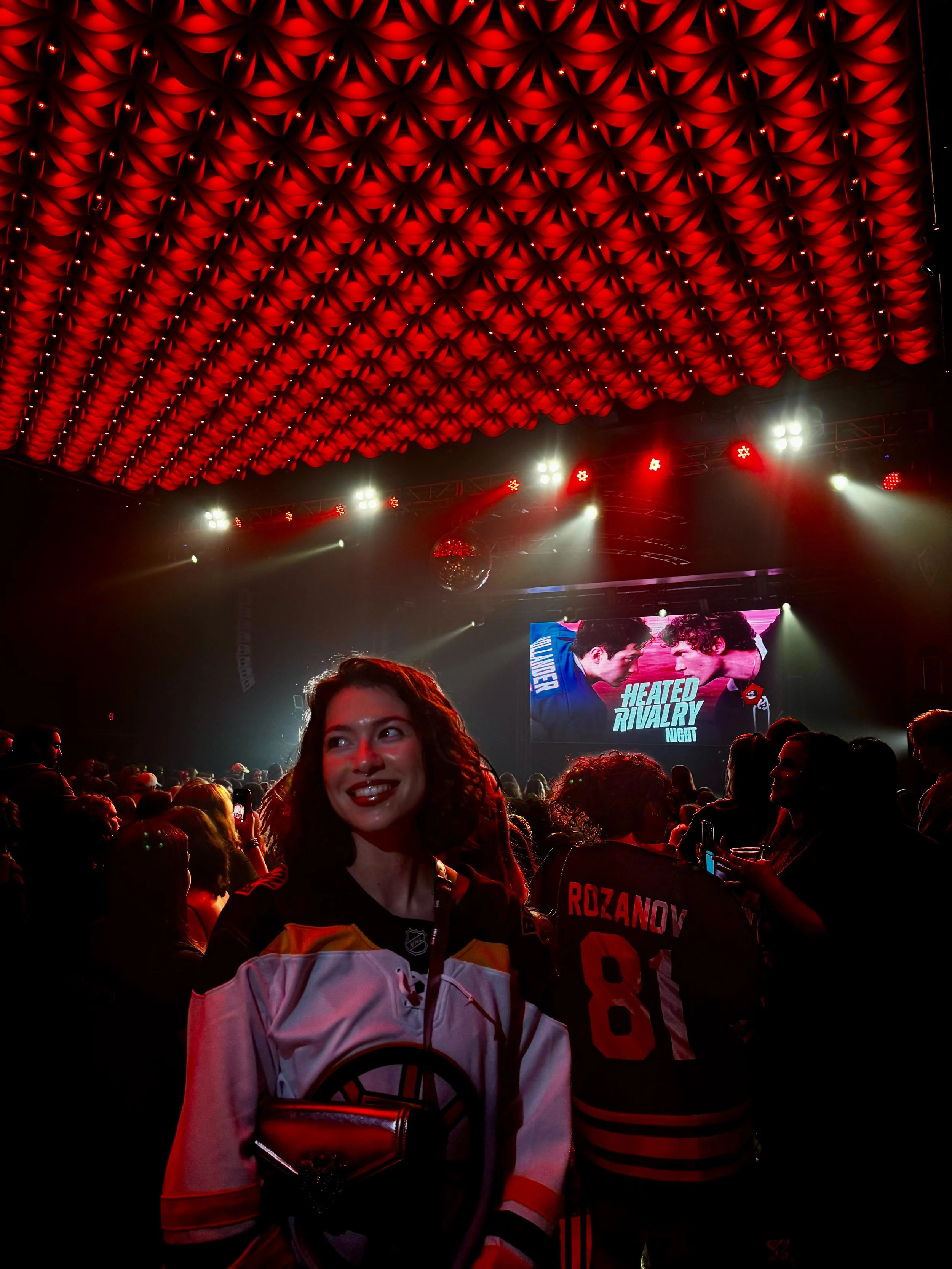 A young woman with curly hair in a hockey jersey smiling in a crowded venue watching a fight or duel on a screen with the words "Heated Rivalry Night" at an event or concert.