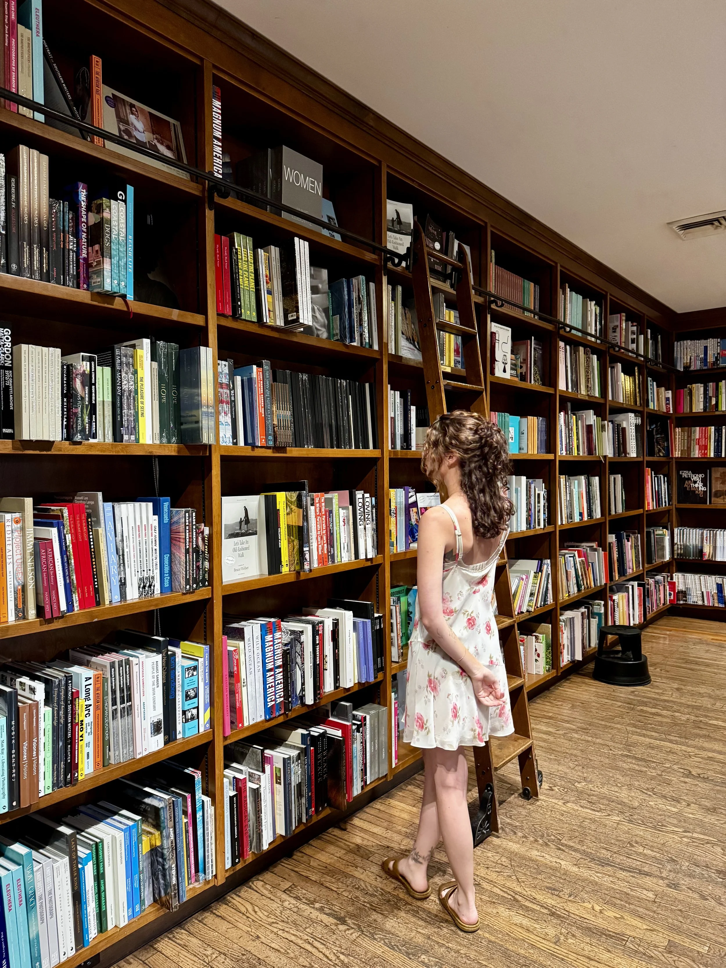 A woman in a floral dress browsing books in a library with wooden shelves and a ladder.