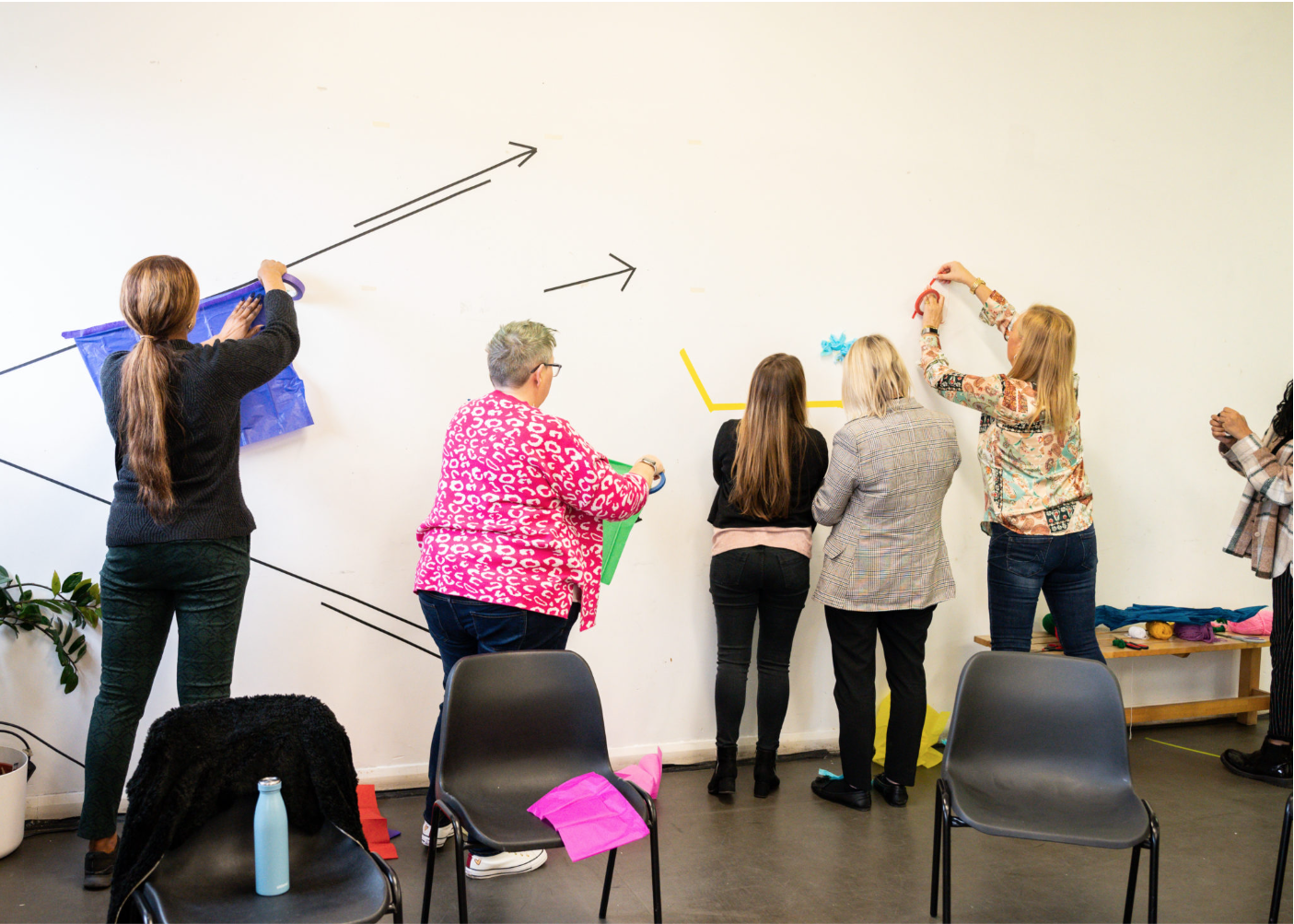 A small group of adult women with their backs turned to the camera sticking paper and cokoured tape shapes across a alrge white wall.