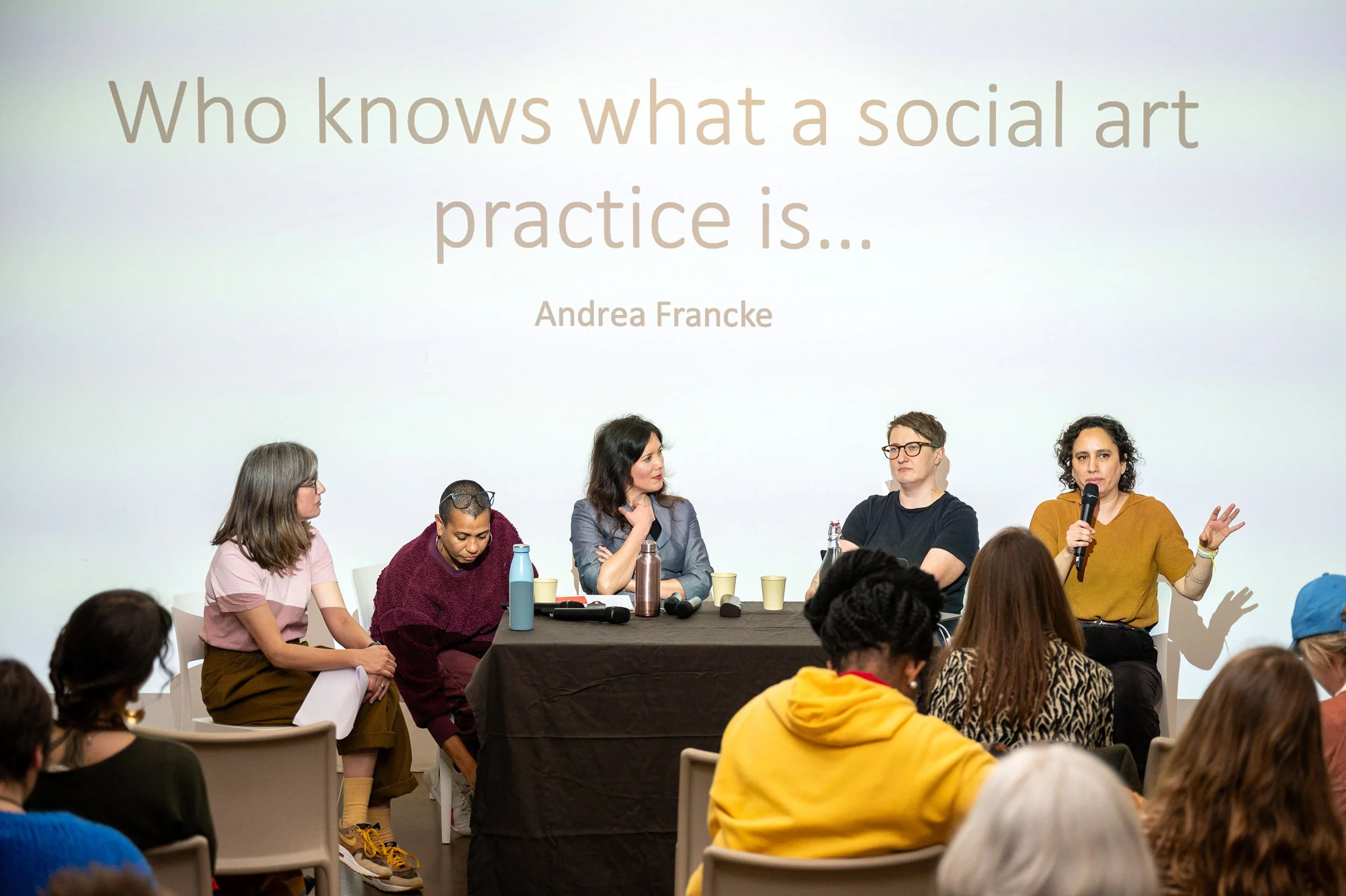 Group of five moment sitting at a table taking part in groups discussion as part of a live event. Audience in foreground. Projected text above them says "Who knows what a social art practice is..."
