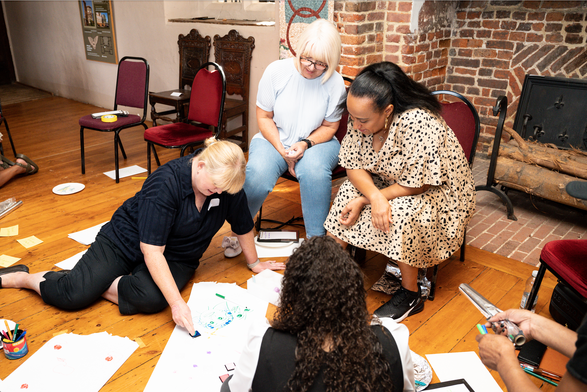 4 adult women crowded around alarge sheet of paper on the floor with one of the group drawing a picture.