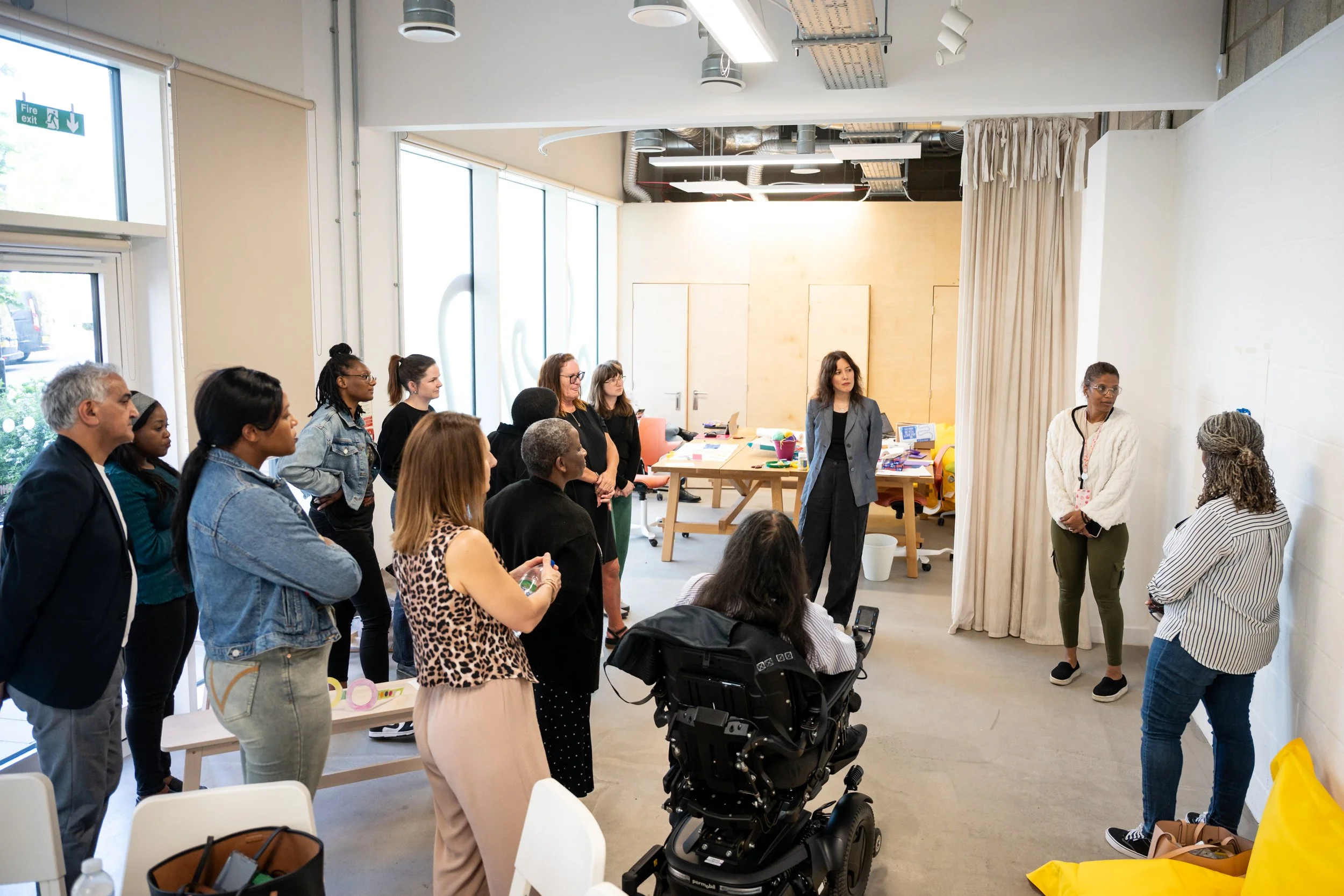Group of 10 adults varying ages, genders, races, ethnicities listening to 2 adult women present something on the wall.
