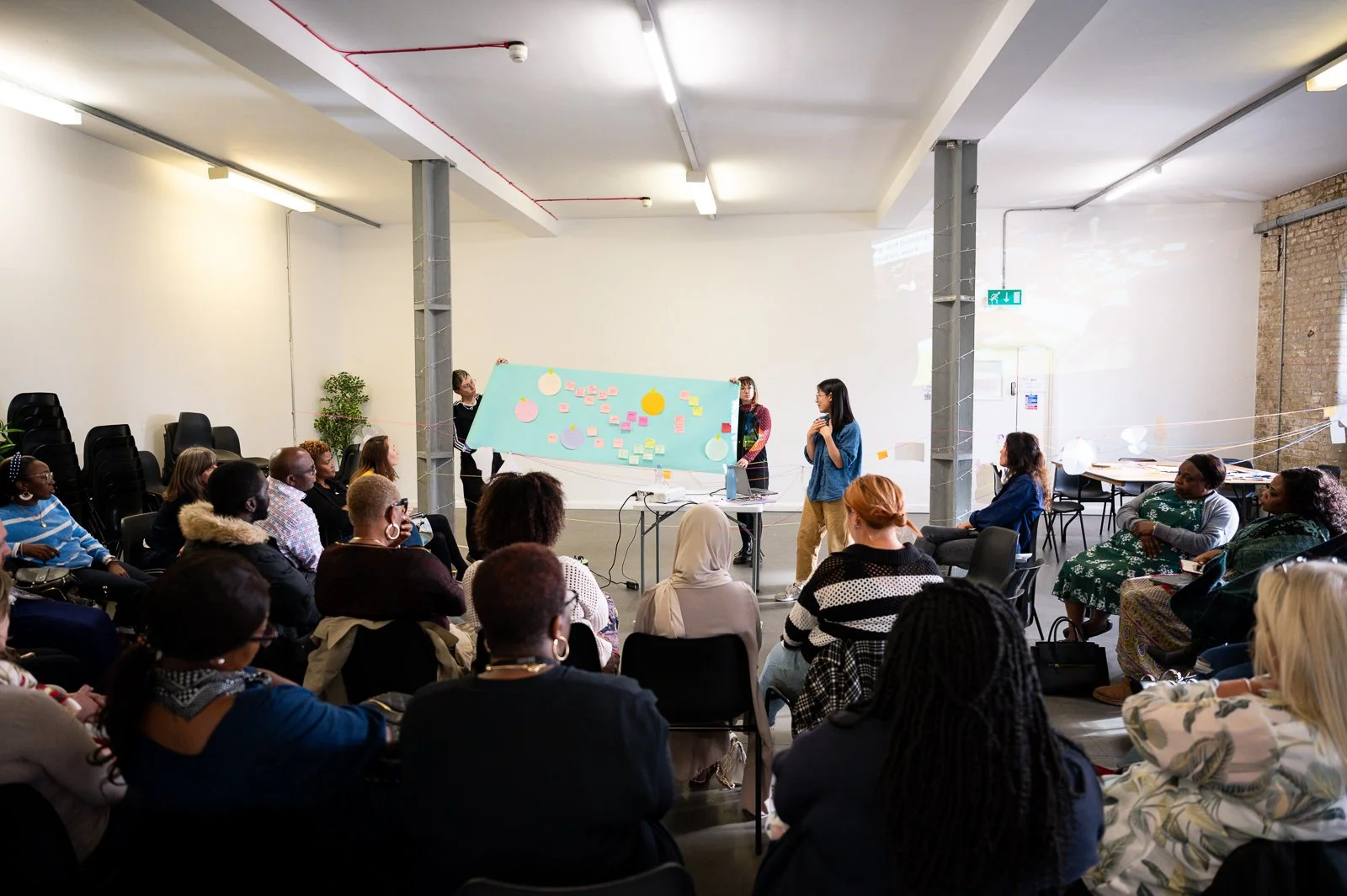 Audience of around 50 people watching 3 adults holding up a long coloured piece of paper with diagrams and drawings whilst talking to the audience.