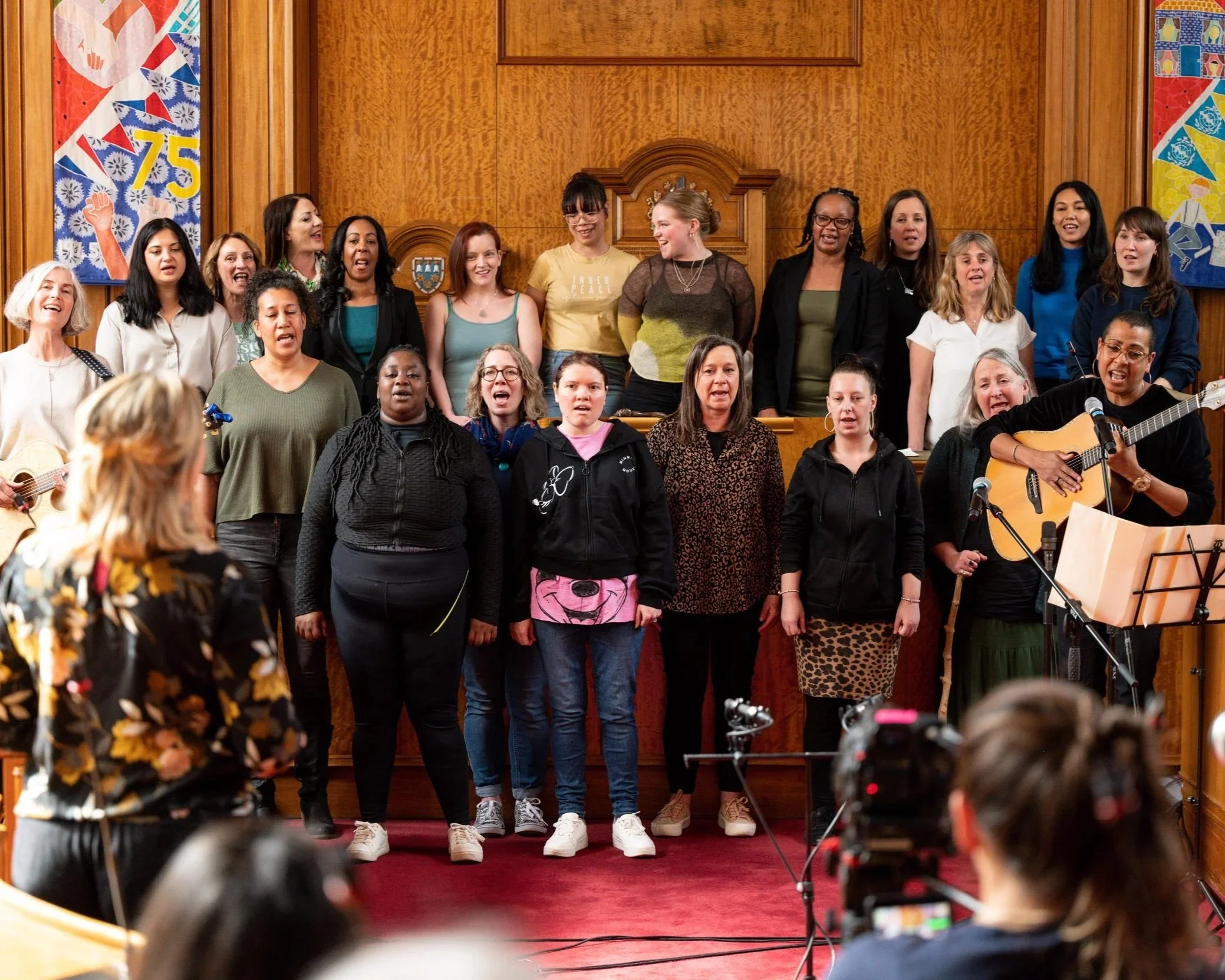 Group of 20 women range in age, race and ethnicity singing whilst Helen Cammock also sings and play the guitar. There are audience members in foreground. The performance is taking place in an oak panelled council chamber.