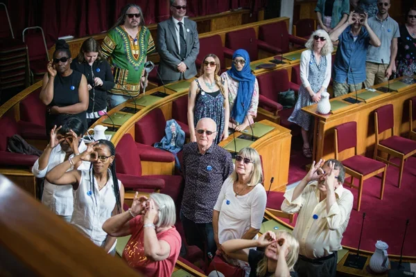 People of varying ages, races and ethncities standing looking towards the camera. Standing in a council assembly room.Some are holding their hands over their eyes like glasses.
