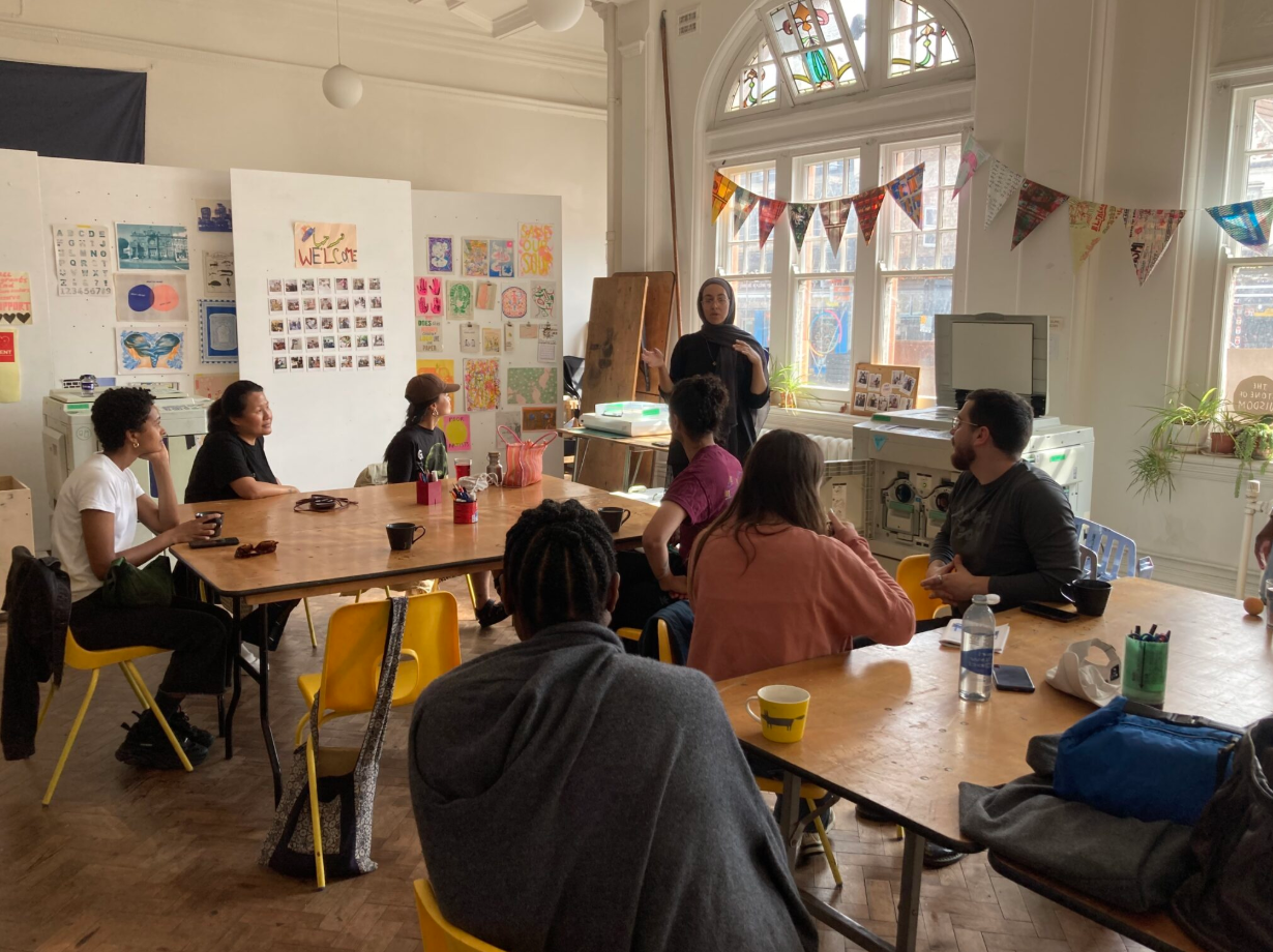 Print studio with 8 adults sitting at tables watching a woman standing and talking to the group.